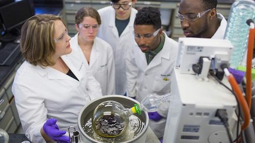 Students in white coats and goggles observing a science experiment